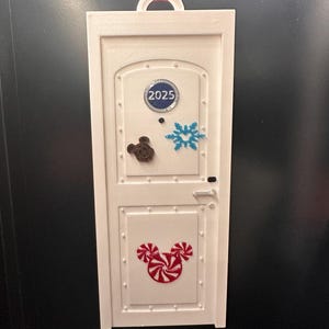 May include: White rectangular ornament shaped like a door, with a red ribbon for hanging. Features a Mickey Mouse candy design, a snowflake, and the year 2025. The ornament has a door handle and rivets.