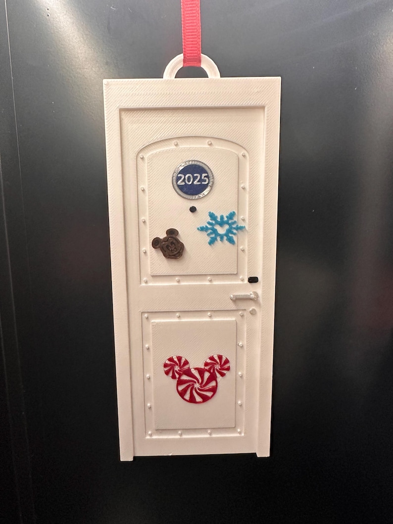 May include: White rectangular ornament shaped like a door, with a red ribbon for hanging. Features a Mickey Mouse candy design, a snowflake, and the year 2025. The ornament has a door handle and rivets.