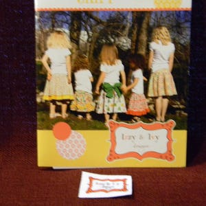 May include: A book cover with a yellow background and a picture of five girls wearing colorful skirts. The book title is "Izzy & Ivy" and the text "Designs" is below the title.