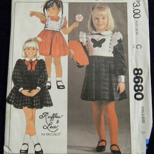 May include: A sewing pattern for a child's dress. The pattern is for a plaid dress with a white collar and a butterfly applique. The pattern is from Ruffles & Lace for McCall's and is size 8680.