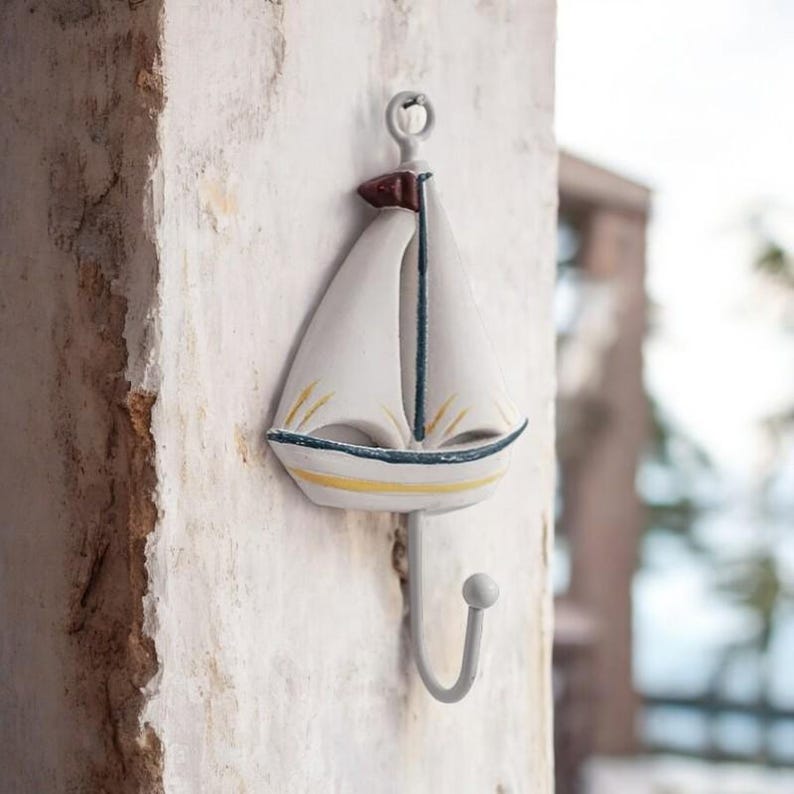 May include: White sailboat shaped metal hook with a yellow stripe and a red flag. The hook is mounted on a white wall.