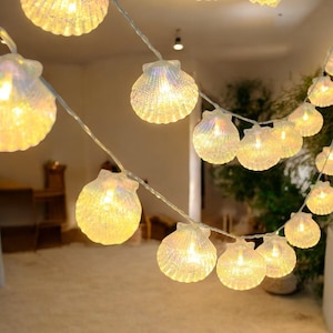 May include: A string of iridescent white seashell-shaped lights with warm white LED bulbs. The lights are strung on a clear wire and are perfect for adding a touch of whimsy to any space.