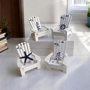 Könnte beinhalten: Vier Miniatur-Adirondack-Stühle aus weißem Holz mit nautischen Dekorationen. Jeder Stuhl hat eine andere Dekoration, darunter ein Leuchtturm, ein Schiffsrad, ein Seestern und ein Segelboot.