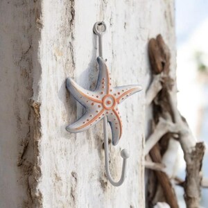 May include: A white metal hook shaped like a starfish with orange accents. The hook is mounted on a white wooden wall.