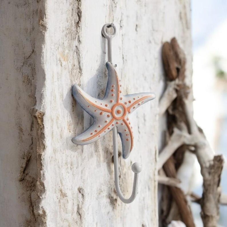 May include: A white metal hook shaped like a starfish with orange accents. The hook is mounted on a white wooden wall.