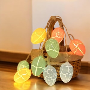 Puede incluir: Una cadena de luces con forma de huevo de Pascua con un acabado craquelado. Las luces son de varios colores, incluyendo amarillo, verde y naranja. Las luces están conectadas entre sí con un cable transparente y cuelgan de una cesta de mimbre.