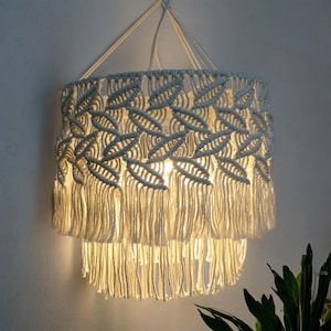 Puede incluir: Una pantalla de lámpara de macramé blanca con un borde de flecos. La pantalla está encendida y emite un brillo cálido.