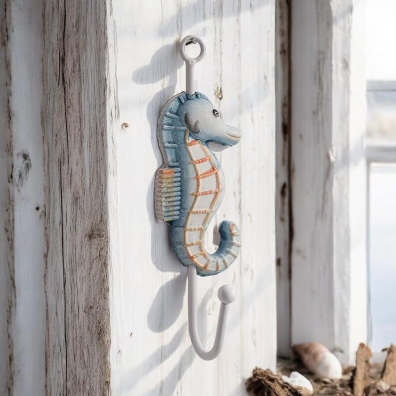 May include: A white metal hook shaped like a blue and orange seahorse. The hook is mounted on a white wooden wall.
