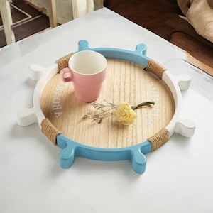 Puede incluir: Una bandeja de madera con forma de rueda de barco con un acabado pintado en azul y blanco. La bandeja tiene un borde de cuerda y las palabras "Ahoy" y "Love" escritas en ella. Una taza rosa y una pequeña flor están en la bandeja.