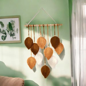 Könnte beinhalten: Ein Wandbehang aus Makrame mit braunen und weißen Blättern. Die Blätter sind aus Garn gefertigt und hängen an einer Holzstange.
