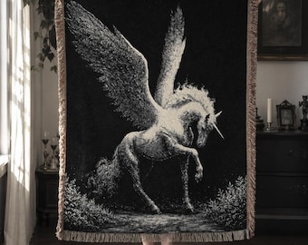 May include: A rectangular woven throw blanket featuring a large, detailed white winged unicorn on a solid black background. The blanket has a textured, fringed border in a light tan color along all four edges, creating a decorative finish.