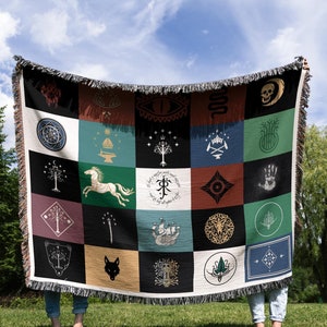 May include: Two people are holding up a tapestry throw blanket with a grid of 25 squares. Each square features a different symbol or design, including a horse, a tree, a hand, and a crown. The blanket is made of a soft, woven fabric and has a fringe border.