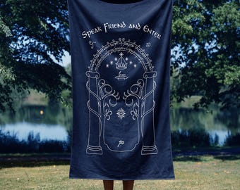 May include: A dark navy blue rectangular towel featuring a white line art graphic of a gateway with two pillars, trees, a crown, and a star. The text "SPEAK FRIEND AND ENTER" is printed in white at the top, with additional script text below it.
