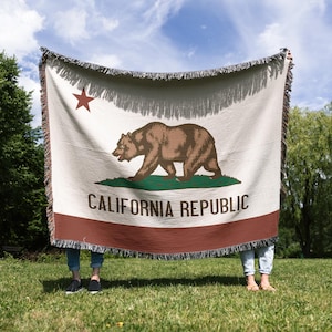 May include: A woven blanket featuring the California state flag design. The flag includes a brown bear, a red star, and the words "California Republic" on a cream-colored background with a brown border and fringe.