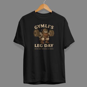May include: A black short-sleeve t-shirt featuring a graphic of a bearded character lifting a barbell. The text reads GYMLI'S LEG DAY and NEVER SKIP THE MINES OF MORIA in a light beige font. The shirt is displayed on a wooden hanger.