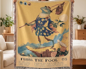 May include: A woven throw blanket featuring a yellow background with a central illustration of a frog wearing a blue floral tunic and hat, carrying a stick with a bag and a white flower. The text reads Flubs, The Fool, Frog Scout, 0/5.