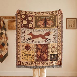 May include: A rectangular woven throw blanket featuring a multi-panel design with a central running fox, a large moth, a heart, a dragonfly, mushrooms, and oak leaves in shades of brown, cream, and dark red with a fringed edge on all sides.
