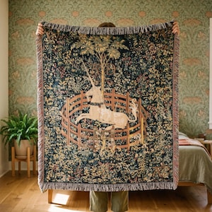 May include: A woven throw blanket featuring a dark blue background with a detailed scene of a white unicorn inside a wooden fence, surrounded by colorful flowers and foliage. The blanket has a fringed edge and a complex, multi-colored tapestry design.