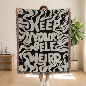 Może przedstawiać: Prostokątny tkany koc z czarnym tłem, jasnoszarymi, falistymi, abstrakcyjnymi wzorami oraz napisem "KEEP YOUR SELF WEIRD" pogrubioną, stylizowaną czcionką. Koc posiada frędzle na górnej i dolnej krawędzi.
