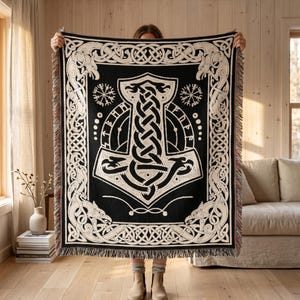 May include: Black throw blanket with a large white Thor's hammer design. The blanket features intricate knotwork and border patterns, with fringe along the edges. The design includes Viking-inspired imagery, perfect for home decor.