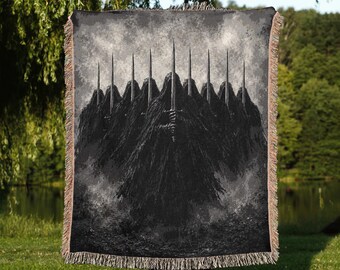 May include: A rectangular woven throw blanket featuring a dark, monochromatic graphic print of nine hooded figures in black cloaks, each holding a vertical sword. The blanket has a textured fringe border along the top and bottom edges.