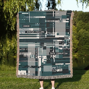 May include: A woven throw blanket featuring a dark teal, black, and white digital glitch pattern. The design includes the text ERROR, SYSTEM FAILURE, and DATA CORRUPTION in a pixelated font. The blanket has a fringed edge on the top and bottom.