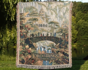 May include: A woven tapestry throw blanket featuring a detailed scene of a stone bridge with figures crossing, set against a backdrop of mountains, waterfalls, and architecture, with the text The Lord of the Rings The Fellowship of the Ring.