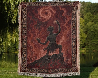 May include: A rectangular woven throw blanket featuring a dark red and black illustration of a creature standing on a rocky outcrop, reaching toward a swirling vortex in the sky. The design is framed by a decorative border with floral motifs.