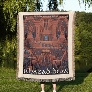 May include: A woven throw blanket featuring a detailed illustration of the mines of Moria. The design includes architectural structures, statues, and a central figure with a beard, with the text Explore the Great Mithril Mines of Moria and Khazad-dûm.