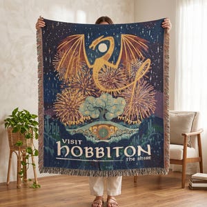Puede incluir: Una manta tejida con un diseño de dragón, fuegos artificiales y el texto "Visit Hobbiton the Shire". La manta tiene un fondo azul oscuro y bordes con flecos. El dragón es dorado y los fuegos artificiales son amarillos y naranjas. Ideal para un sofá.