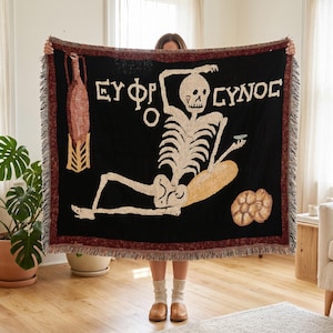 May include: A black woven throw blanket featuring a cream-colored skeleton graphic, a brown amphora, and two round bread loaves. The text "ΕΥΦΡΟΣΥΝΟΣ" is printed in white above the skeleton. The blanket has a dark red border and fringed edges.