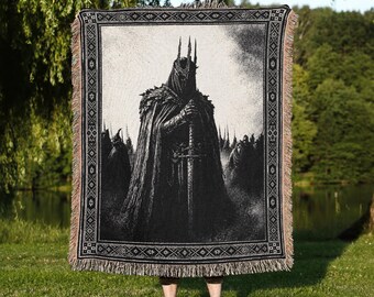 May include: A rectangular woven throw blanket featuring a black and white graphic illustration of a dark armored figure wearing a spiked crown and holding a long sword, surrounded by shadowy figures, with a decorative geometric border and fringe.