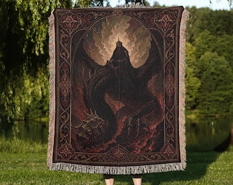 May include: A rectangular woven throw blanket featuring a dark, detailed illustration of a winged, horned creature standing amidst flames and smoke. The design is framed by an ornate, decorative border with fringe along the top and bottom edges.