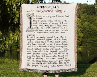 May include: A rectangular woven throw blanket featuring the printed text of Chapter One, An Unexpected Party, from The Hobbit. The off-white fabric displays dark brown lettering, a decorative initial letter, and fringed edges on the top and bottom.
