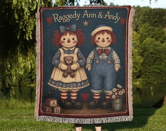 May include: A woven throw blanket featuring the text Raggedy Ann & Andy with illustrations of the two characters. The design includes a dark blue background, red hearts, alphabet blocks, a teddy bear, and a bucket of flowers with fringed edges.