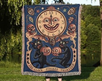 May include: A rectangular woven throw blanket featuring a dark blue background with a central clock face showing the number thirteen. The design includes two black cats holding pumpkins, skulls, a bat, and the text THE CLOCK STRIKES THIRTEEN.