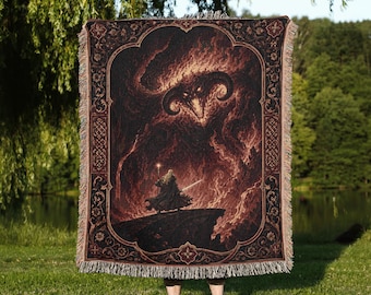 May include: A rectangular woven throw blanket featuring a dark, fiery illustration of a horned demon looming over a cloaked figure holding a staff. The design is framed by an ornate, dark border with intricate patterns and has a fringed edge.