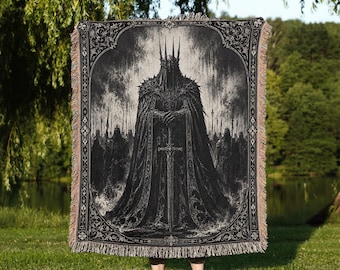 May include: A rectangular woven throw blanket featuring a black and white illustration of a crowned figure in dark armor holding a long sword. The design is framed by an ornate border and finished with fringed edges on the top and bottom sides.