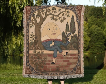 May include: A woven throw blanket featuring a detailed illustration of Humpty Dumpty sitting on a brick wall. The character wears a blue suit with a red patterned cravat and light-colored shoes, holding a book under a tree with a dark border.
