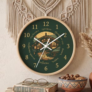 May include: A round wall clock with a light wood frame and a dark green face. The clock features gold numbers and text reading Elevensies, Luncheon, Afternoon Tea, Dinner, Supper, Breakfast, and Second Breakfast surrounding a central illustration.