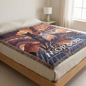 May include: A woven throw blanket featuring a dark, mountainous landscape with a tall tower topped by a glowing eye. The text MORDOR the land of shadow is printed in white capital letters. The blanket has a fringed edge and a multi-colored design.