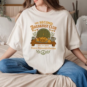 Puede incluir: Una camiseta blanca con un diseño gráfico de una puerta con un cartel que dice "The Second Breakfast Club Est. TA 2880 Peace, Pipeweed & Pastries". El gráfico también incluye una pipa, pan y tazas.