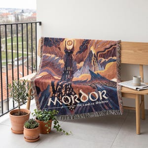 May include: A woven throw blanket featuring a detailed illustration of a dark tower with a glowing eye at the top, a volcanic mountain, and jagged terrain. The text "MORDOR the Land of Shadow" is printed in white at the bottom of the design.