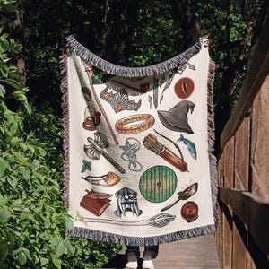 May include: A cream-colored woven throw blanket featuring a variety of illustrated fantasy items, including a golden ring with script, a pointed hat, a green round door, a battle axe, a quiver of arrows, a horn, a helmet, and a staff with fringe edges.
