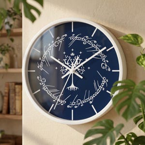 May include: A round wall clock with a white frame and a dark blue face. The clock features a white graphic of a stylised tree centred within a circular border of white Elvish script. It has white hour, minute, and second hands and white tick marks.