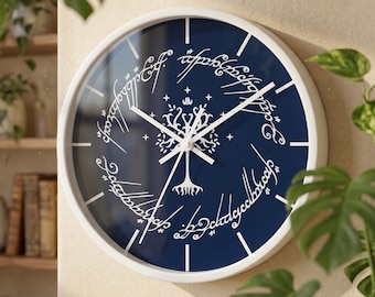 Puede incluir: Un reloj de pared redondo con marco blanco y esfera azul oscuro. El reloj presenta un gráfico blanco de un árbol estilizado centrado dentro de un borde circular de escritura élfica blanca. Tiene manecillas blancas y marcas de hora blancas.
