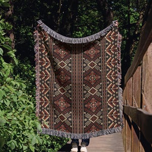 Op de afbeelding: Een rechthoekige geweven plaid met een donkerbruine en zwarte achtergrond en een herhalend geometrisch patroon in beige, crème en roestoranje. De deken is voorzien van decoratieve franjes aan de boven- en onderkant met een symmetrisch ontwerp.