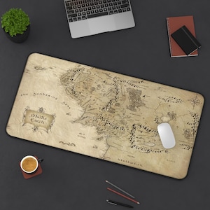 May include: A rectangular desk mouse pad featuring a map of Middle Earth in a vintage style. The map is in shades of brown and tan, with black text and illustrations. A white mouse rests on the pad, with a laptop and other office supplies in the background.