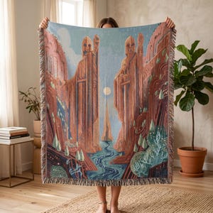 May include: Woven throw blanket featuring a landscape scene with two large statues, a river, and mountains in shades of red, blue, and green. The blanket has a fringed edge and is held up by a person.