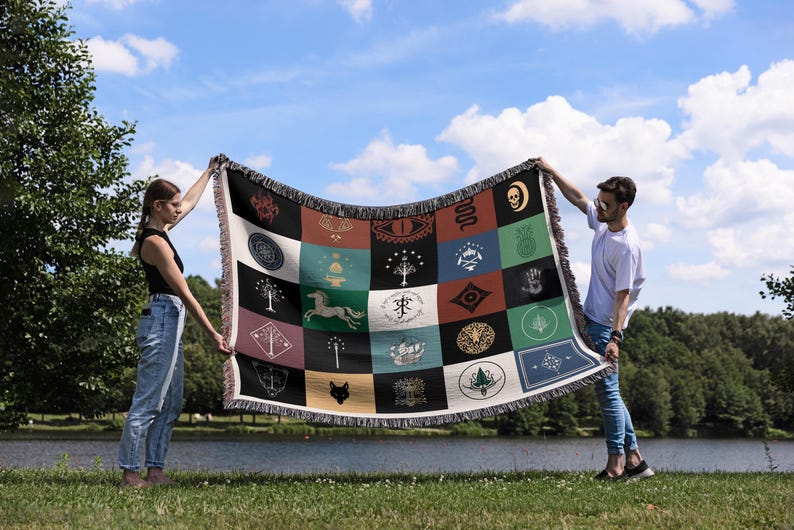 Lord of the Rings Fantasy emblems throw blanket held by pair outdoors, woven patchwork design with magical symbols, decorative fringe by lakeside